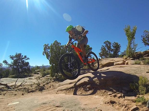 A mountain biker in a green helmet performs a jump on a rocky trail surrounded by trees under a clear blue sky. The biker is wearing a backpack and is mid-air above the ground. Little Canyon mountain bike trail.