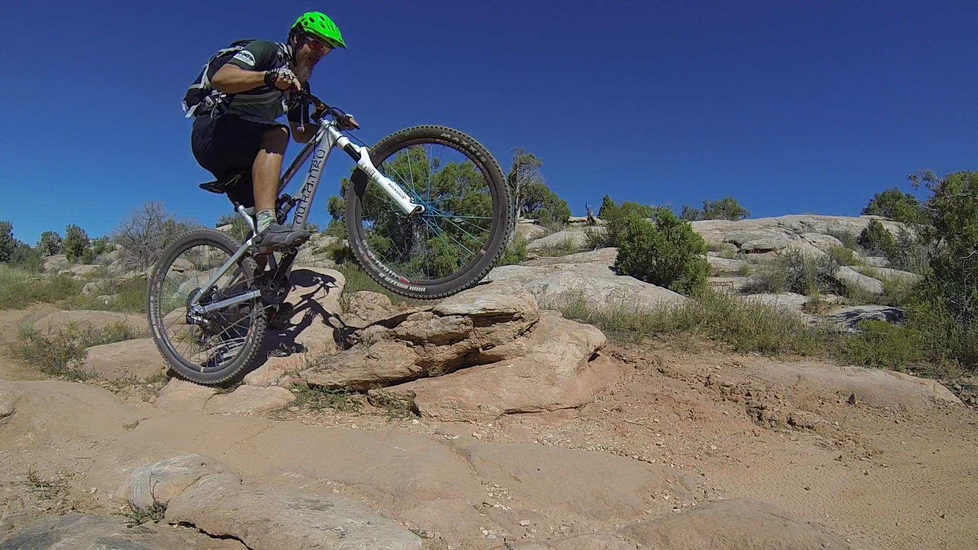 Mountain biker performing a jump over a large rock on a rugged trail, surrounded by shrubs and boulders under a clear blue sky. Moab Brand Trails mountain bike trail.
