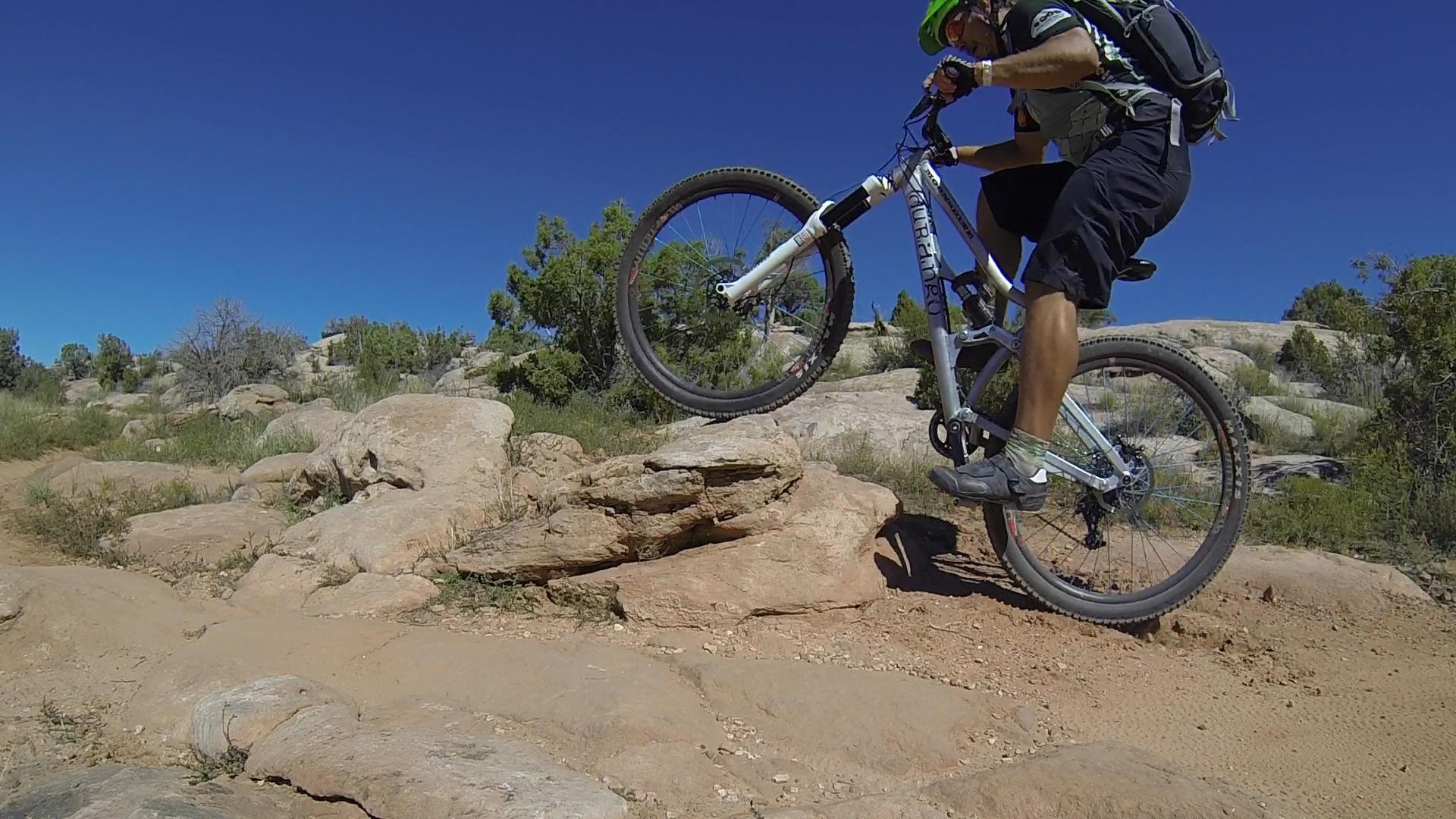 A mountain biker performing a jump over a rocky terrain, with bright blue skies and greenery in the background. The cyclist is wearing a helmet and riding a mountain bike with one wheel raised off the ground, showcasing an action-packed moment in an outdoor setting. Moab Brand Trails mountain bike trail.
