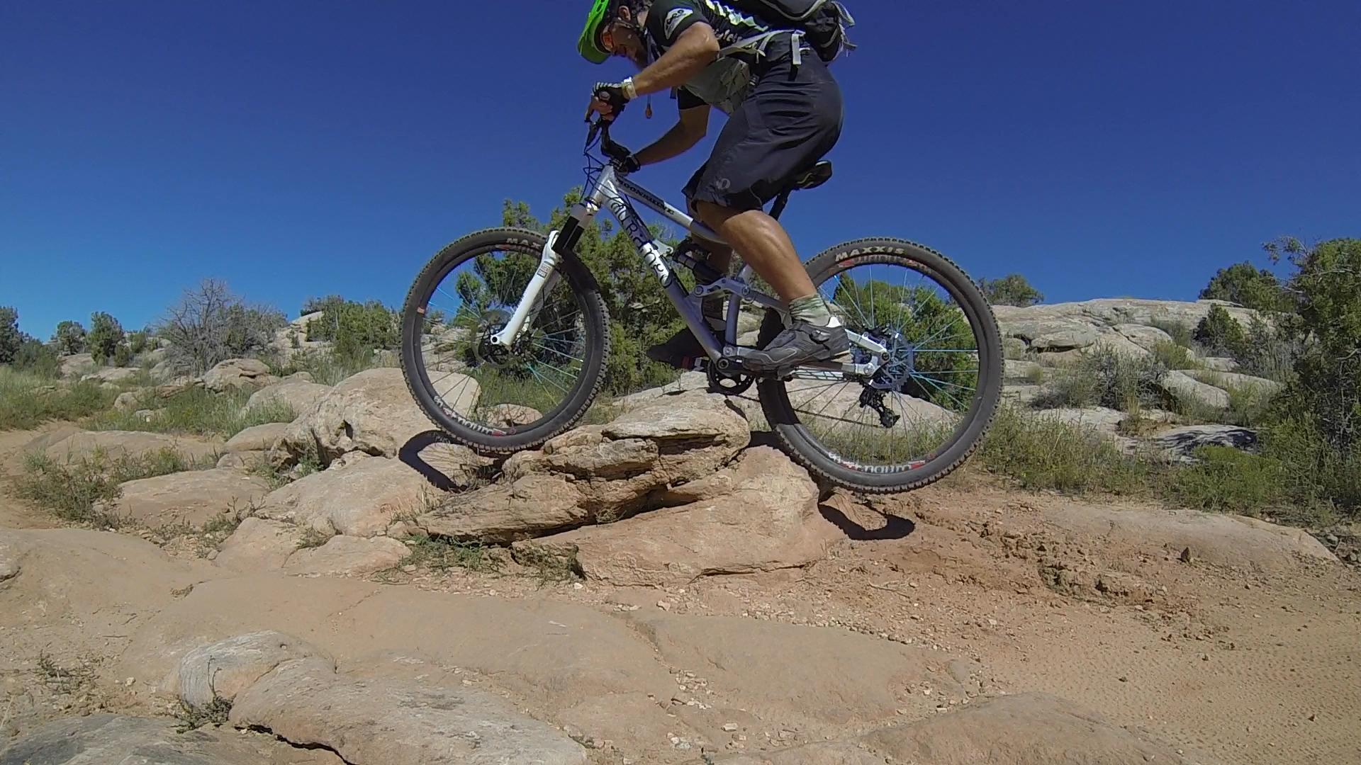 A mountain biker launching off a rock on a dirt trail, with blue skies and greenery in the background. The rider is wearing a helmet and riding a white mountain bike, showcasing an action-packed moment in outdoor cycling. Moab Brand Trails mountain bike trail.