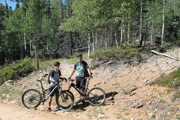 Two cyclists, a child and an adult, stand beside their mountain bikes on a dirt trail surrounded by trees. The background features green foliage and aspen trees, indicating a forested area. The sun shines brightly, suggesting a clear day.