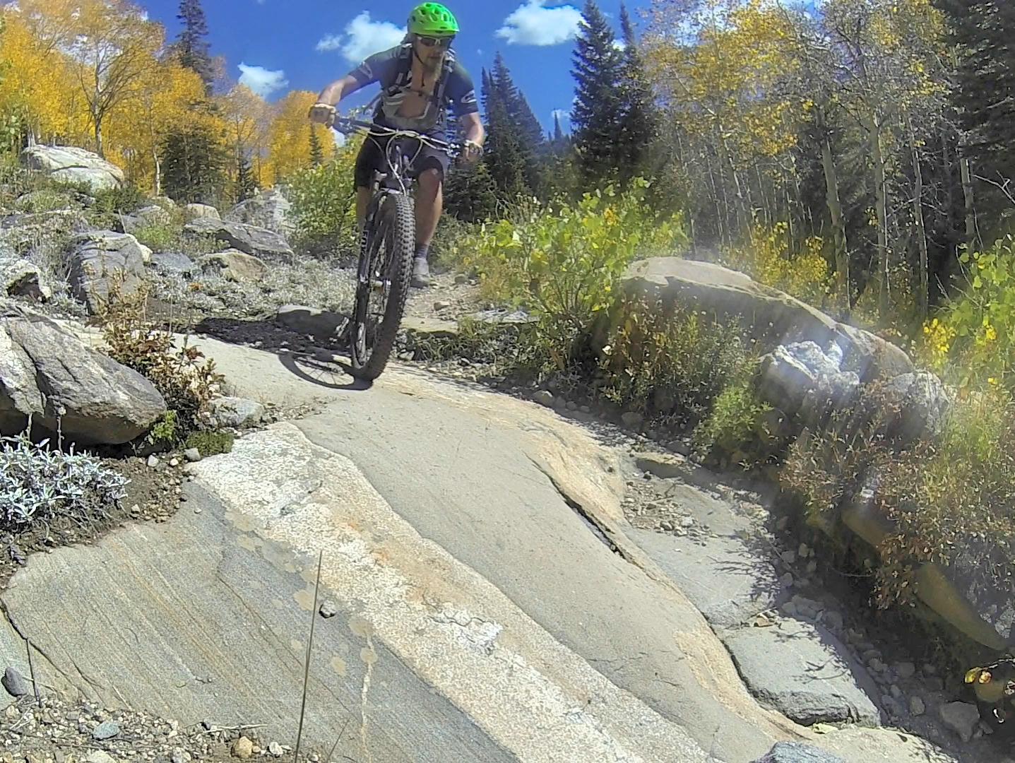 A mountain biker navigating a rocky trail surrounded by autumn foliage and tall trees under a blue sky with fluffy clouds. Fish Creek Falls mountain bike trail.