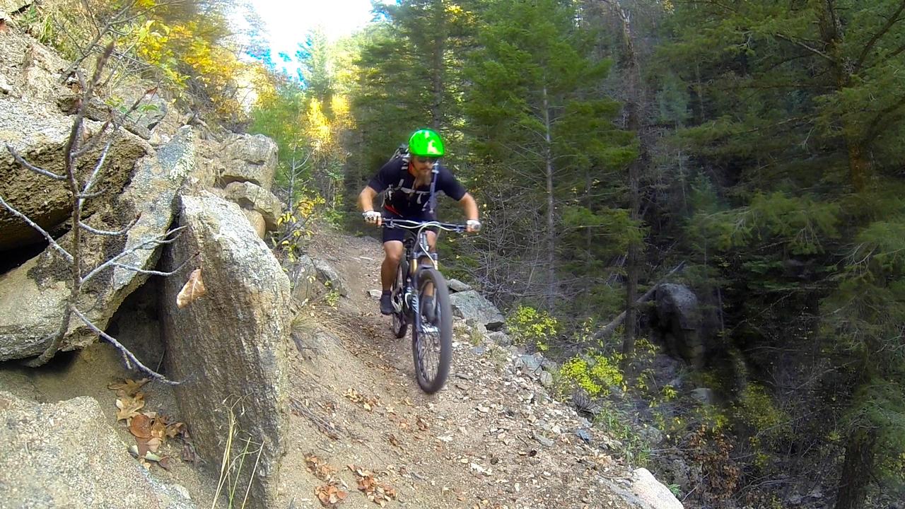 A mountain biker with a green helmet rides along a narrow dirt trail surrounded by rocks and trees in a forested area. Hot Springs Trail mountain bike trail.