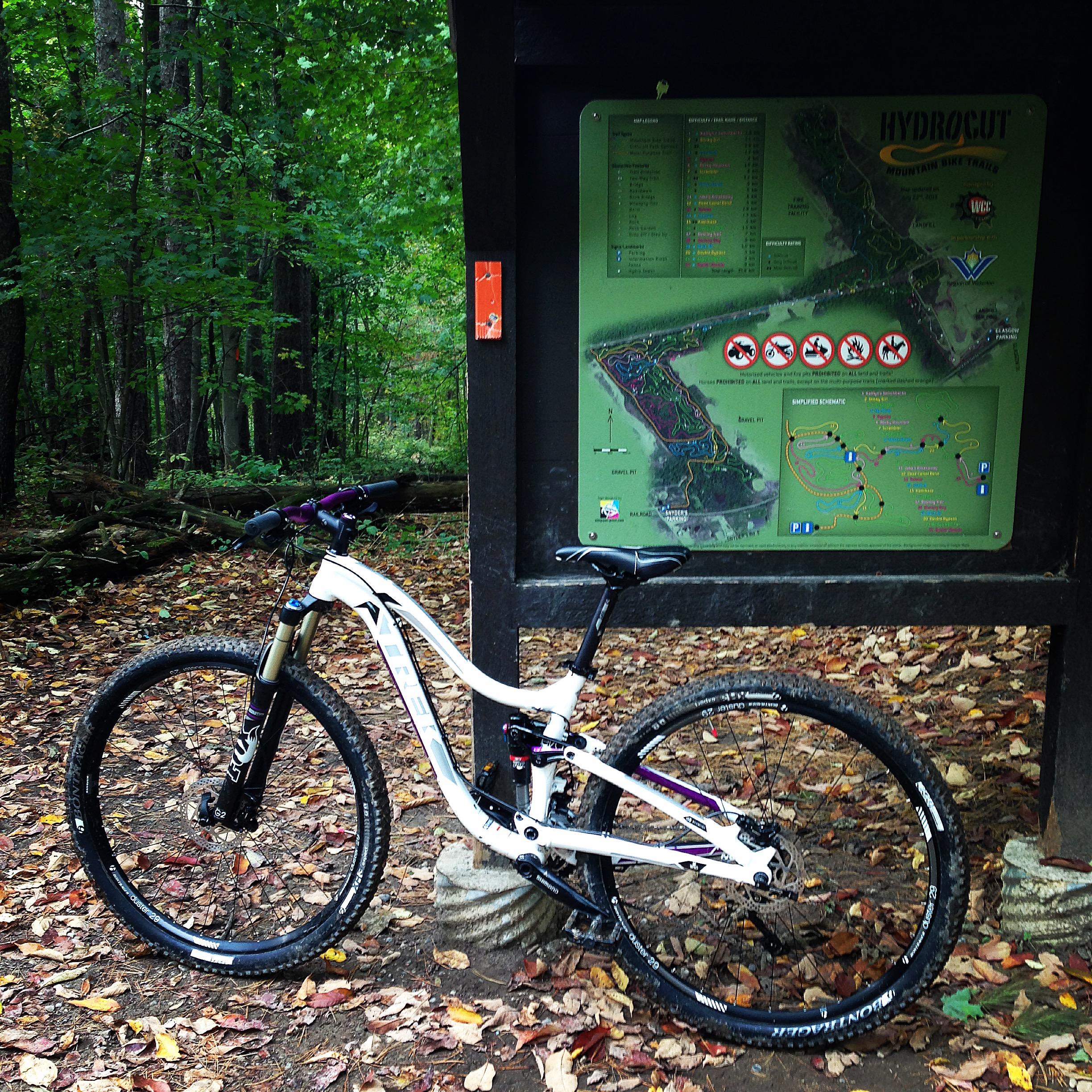A mountain bike resting on a dirt path covered in fallen leaves, next to a trail map sign for "Hydrocut." The sign features various colored trails and symbols indicating rules and facilities, surrounded by a lush, green forest. Hydrocut mountain bike trail.
