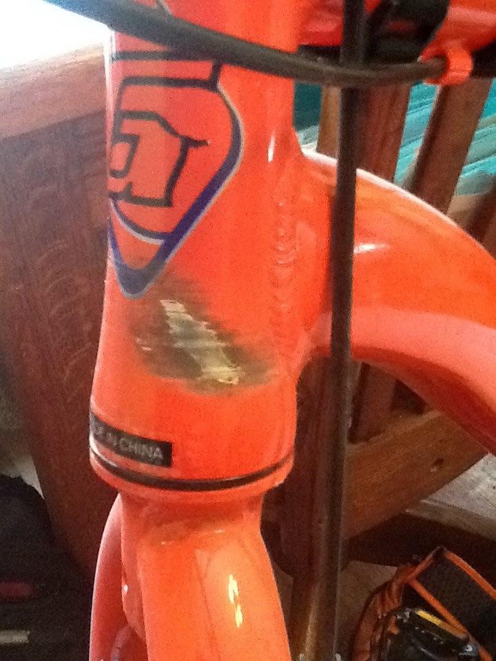 Gravity G29FS: Close-up of an orange bicycle frame featuring a logo on the neck area, with some wear and scratches visible. The frame is attached to a bicycle stand, with wooden background elements partially visible.