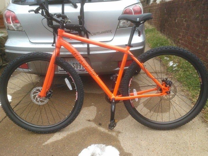Gravity G29FS: A bright orange mountain bike is parked next to a silver SUV. The bike features thick tires and disc brakes, with a visible bike rack on the back of the vehicle. The ground is partially wet, indicating recent rain or snow.