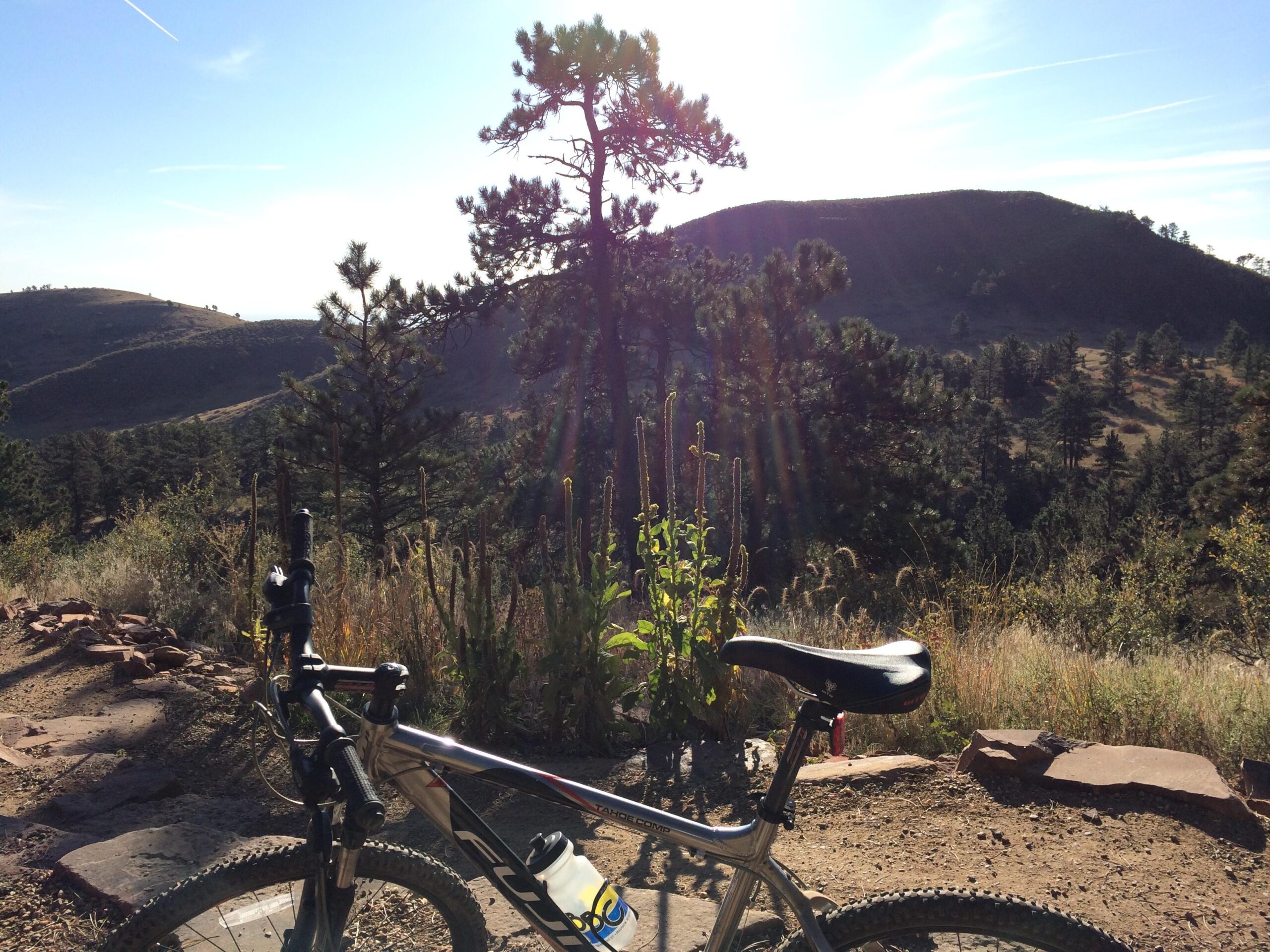 A mountain bike positioned on a rocky trail, with a scenic view of rolling hills and tall pine trees in the background, under a clear blue sky with the sun shining brightly. Heil Valley Ranch mountain bike trail.