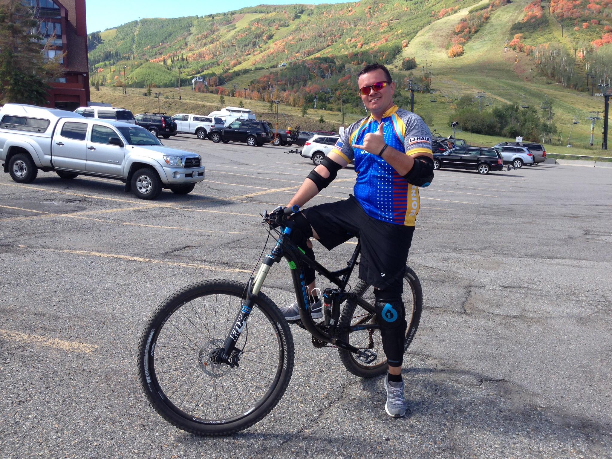 Trek Remedy 8 29: A mountain biker posing with a thumbs-up while sitting on his bike in a parking lot, surrounded by vehicles. He is wearing a colorful cycling jersey and protective gear, with a scenic hillside featuring autumn foliage in the background.