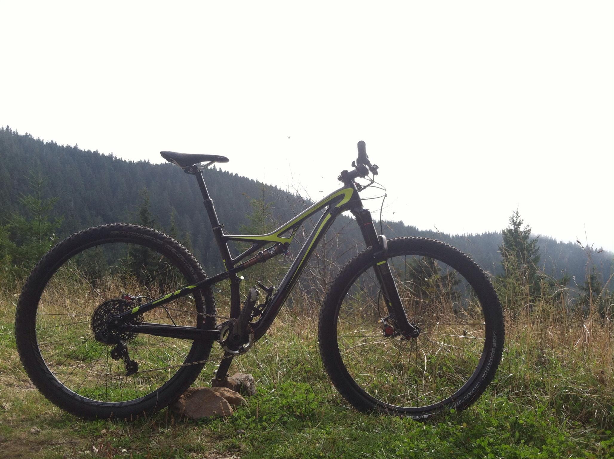 Specialized Camber Expert Carbon EVO 29: A close-up view of a black mountain bike with green accents, positioned on a grassy area with rocky terrain. In the background, dense green trees and a misty mountain landscape are visible, under a cloudy sky. The bike is angled to showcase its features, such as the wheels, frame, and handlebars.