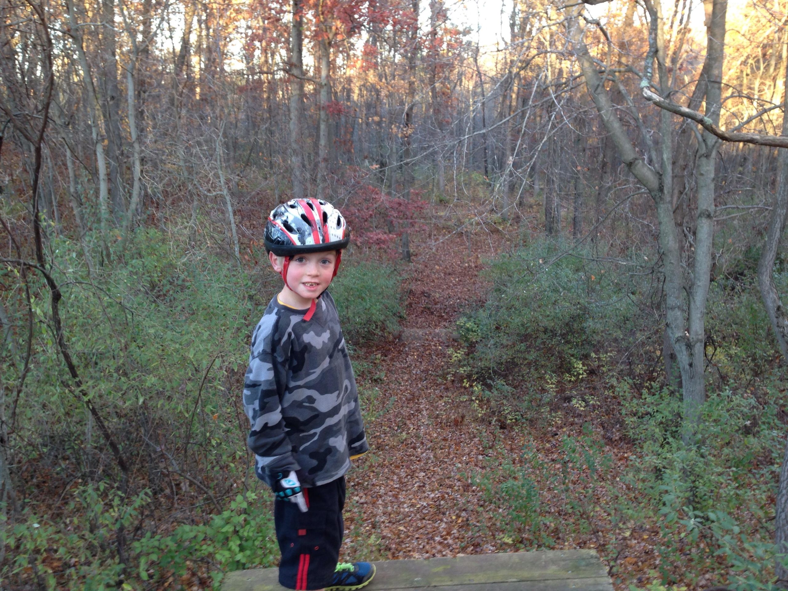 A young child wearing a helmet and a camouflage sweatshirt stands on a wooden platform in a wooded area. The background features trees with autumn foliage and a winding dirt path covered in fallen leaves. Cannonsburg Ski Area mountain bike trail.