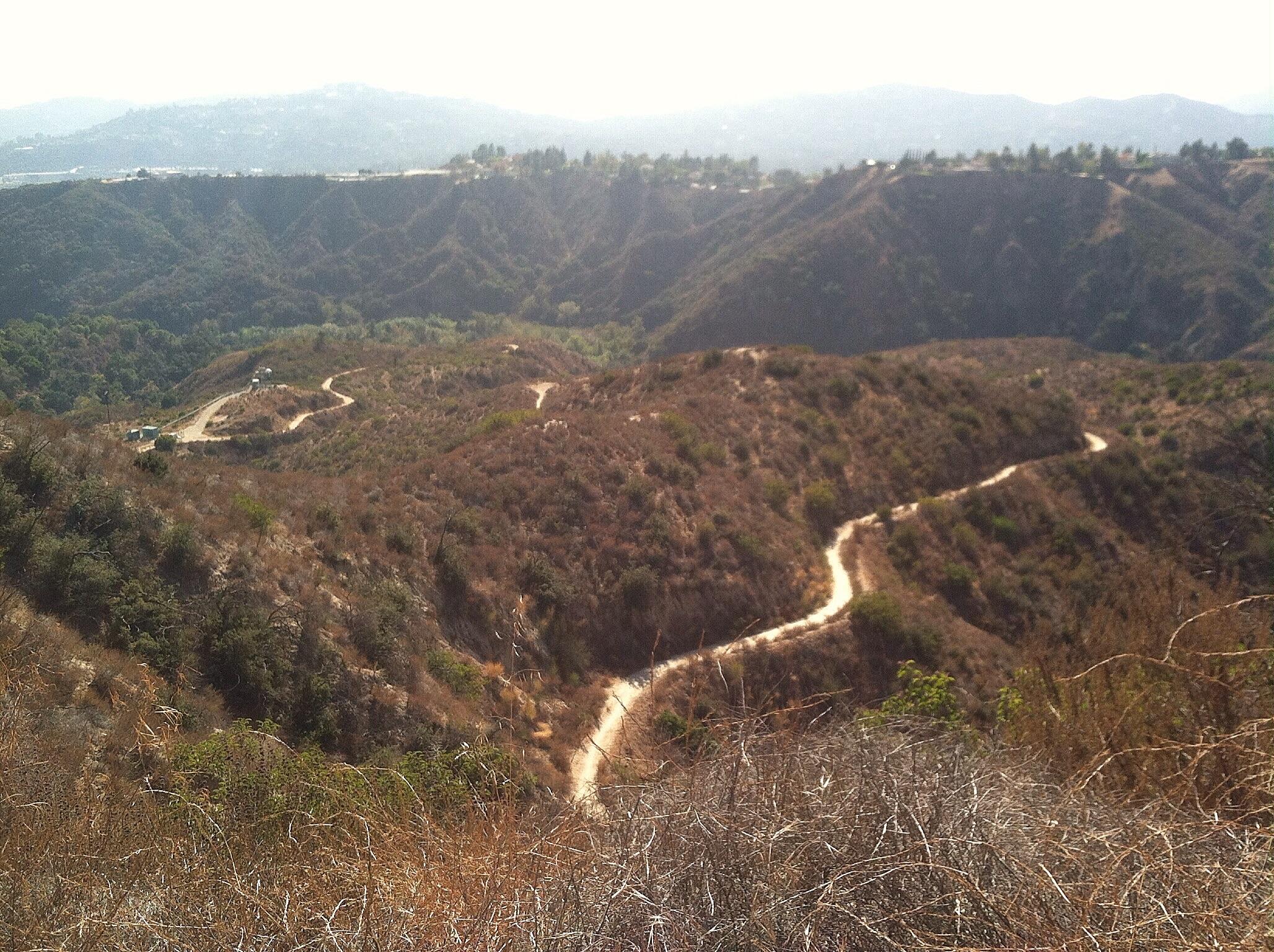 A scenic view of rolling hills and winding dirt paths in a dry, mountainous landscape under a hazy sky. Vegetation is sparse with patches of green amid the brown, sunlit terrain. El Prieto mountain bike trail.