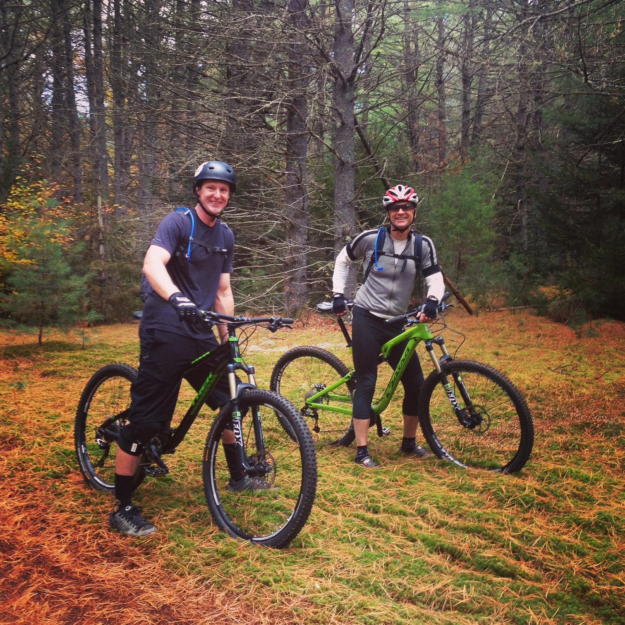 Two men smiling and posing with mountain bikes in a forested area, surrounded by tall trees and autumn foliage. They are wearing helmets and athletic clothing, standing on a bed of pine needles and grass. Kingdom Trails mountain bike trail.