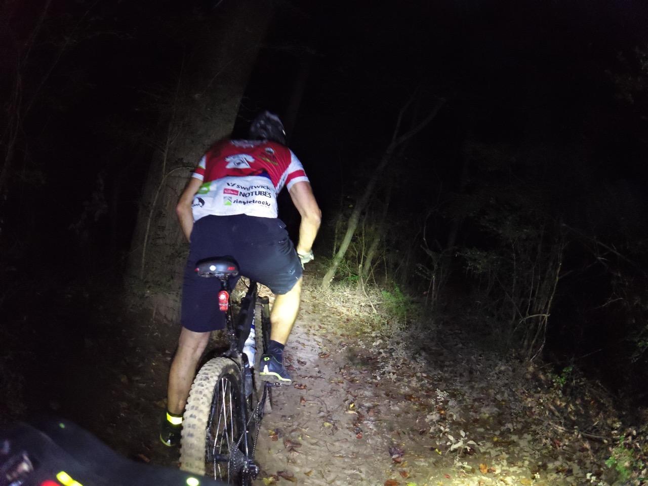 A mountain biker riding on a dimly lit trail at night, surrounded by trees and fallen leaves. The rider is wearing a red and white shirt and black shorts, and the bike is equipped with lights for visibility. Ira B Melton mountain bike trail.