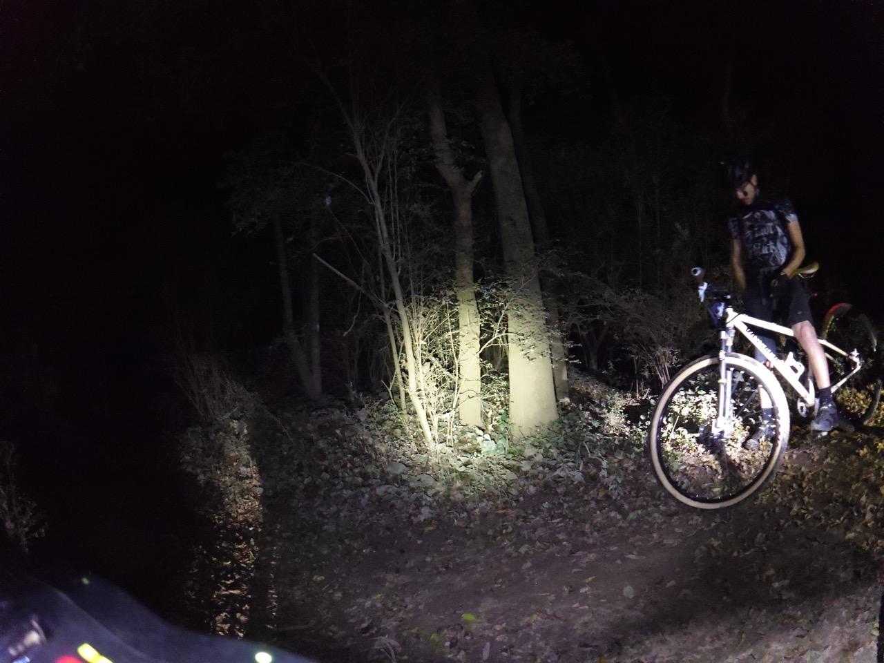 A cyclist in a dark forest at night, illuminated by a bike light. The rider is standing next to a white mountain bike on a dirt path surrounded by trees and fallen leaves. Ira B Melton mountain bike trail.