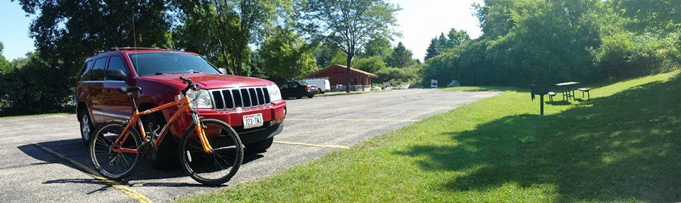 Gary Fisher Advance: A bright sunny day in a parking area featuring a red SUV parked next to an orange bicycle. In the background, there is a picnic area with a red building and picnic tables surrounded by greenery. The parking lot is mostly empty with well-maintained grass on the side.
