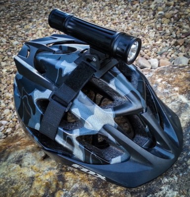 A black and gray camo-patterned bicycle helmet with a mounted flashlight on top, resting on a rocky surface.