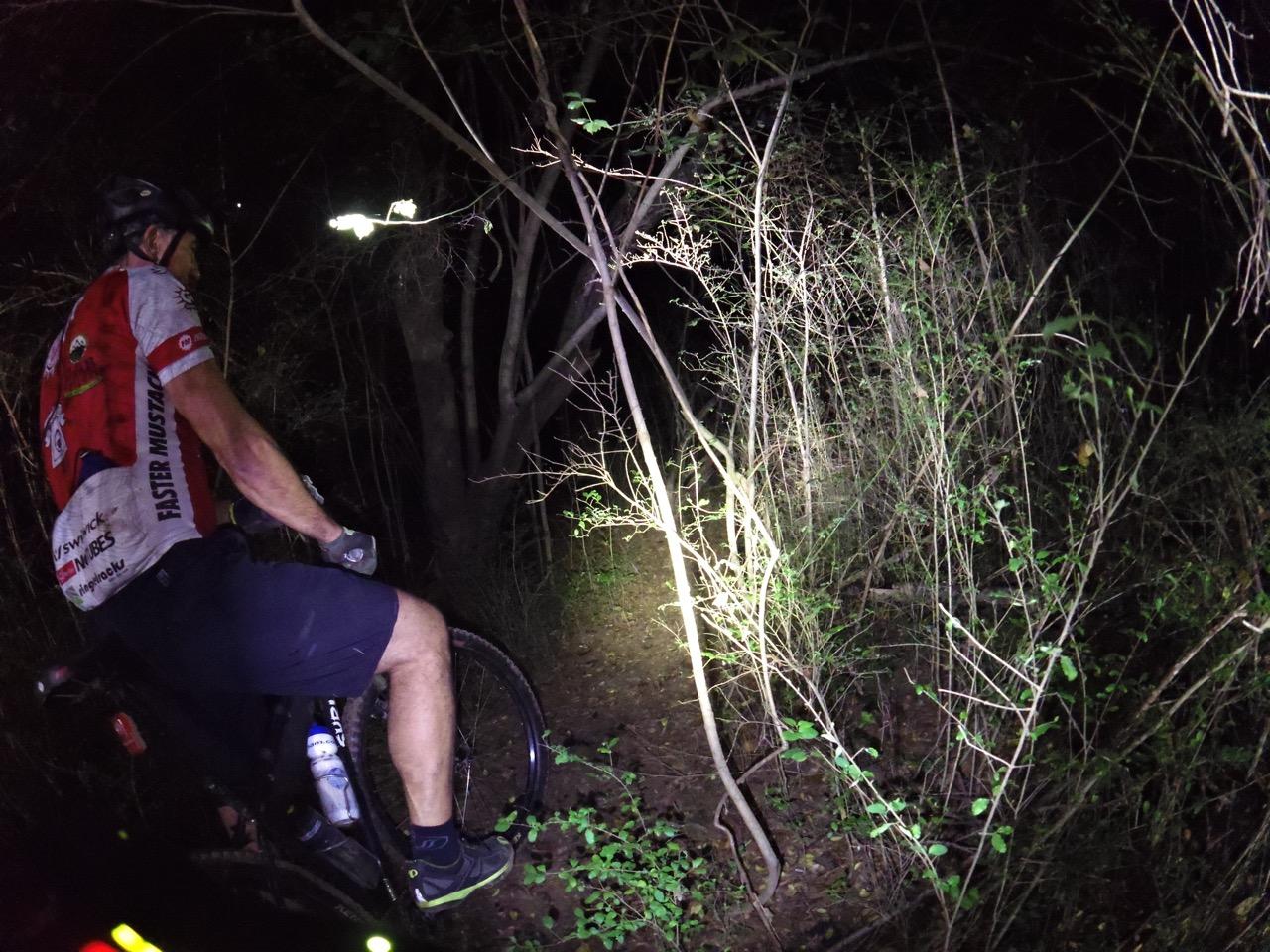 A cyclist dressed in a red and white jersey navigates a dark forest trail at night, using a bike-mounted light to illuminate the path ahead. The surroundings are dense with trees and underbrush, creating a rugged natural environment. Waterworks / Mason Mill mountain bike trail.