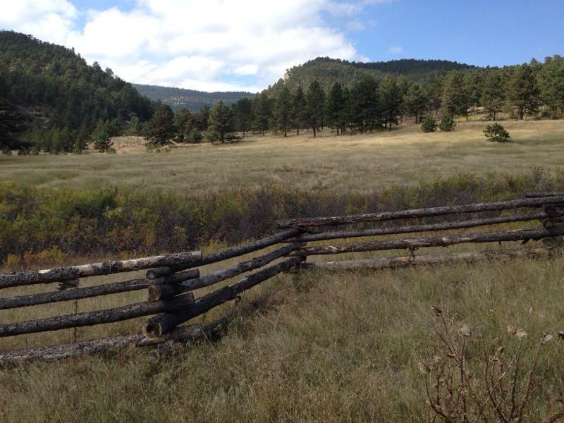 A scenic view of a green meadow bordered by a rustic wooden fence, surrounded by pine trees and rolling hills under a partly cloudy sky. Heil Valley Ranch mountain bike trail.