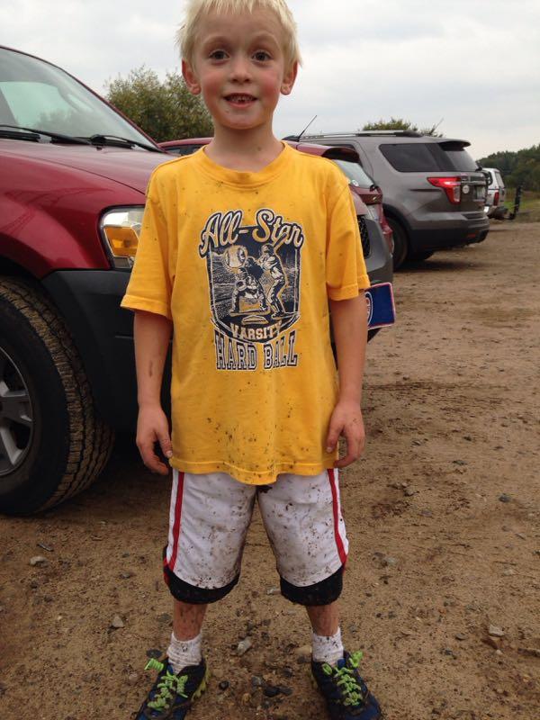 A smiling young boy with messy, dirt-covered clothes stands in a gravel parking area. He is wearing a yellow T-shirt with a sports graphic and red and white shorts. His shoes are also dirty, and he has light blonde hair. Several parked vehicles are visible in the background under an overcast sky. Merrell Trail mountain bike trail.