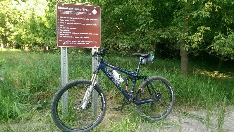 A mountain bike leaning against a trail sign in a grassy area, with trees in the background. The sign provides guidelines for riding on the mountain bike trail. Kiwanis - Mankato mountain bike trail.
