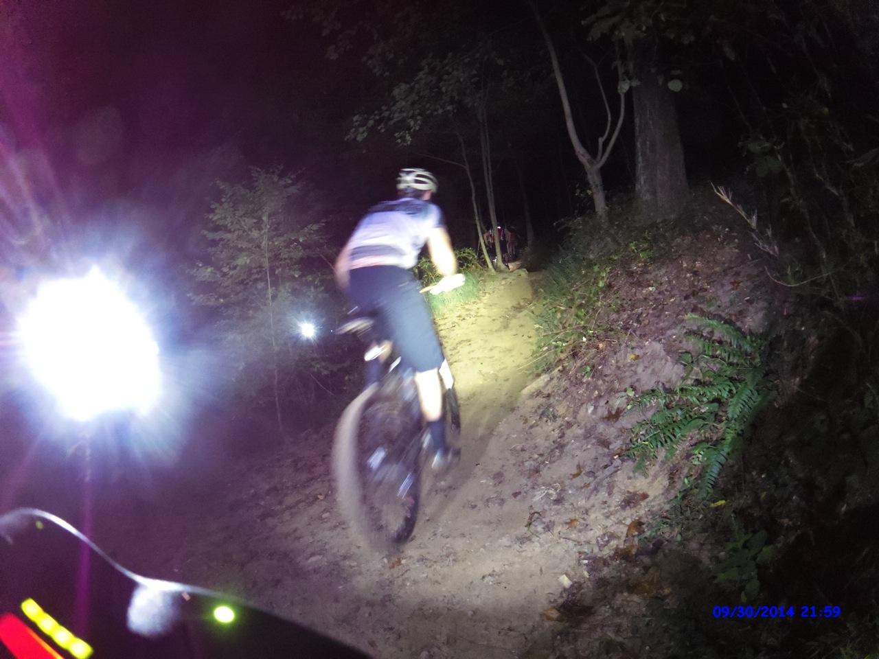 A cyclist riding a mountain bike on a dirt trail at night, illuminated by bright bike lights. The scene shows a dimly lit forest with trees and ferns on either side of the trail. The cyclist is blurred in motion, suggesting speed, while additional lights in the background indicate other riders. Morningside Nature Preserve mountain bike trail.
