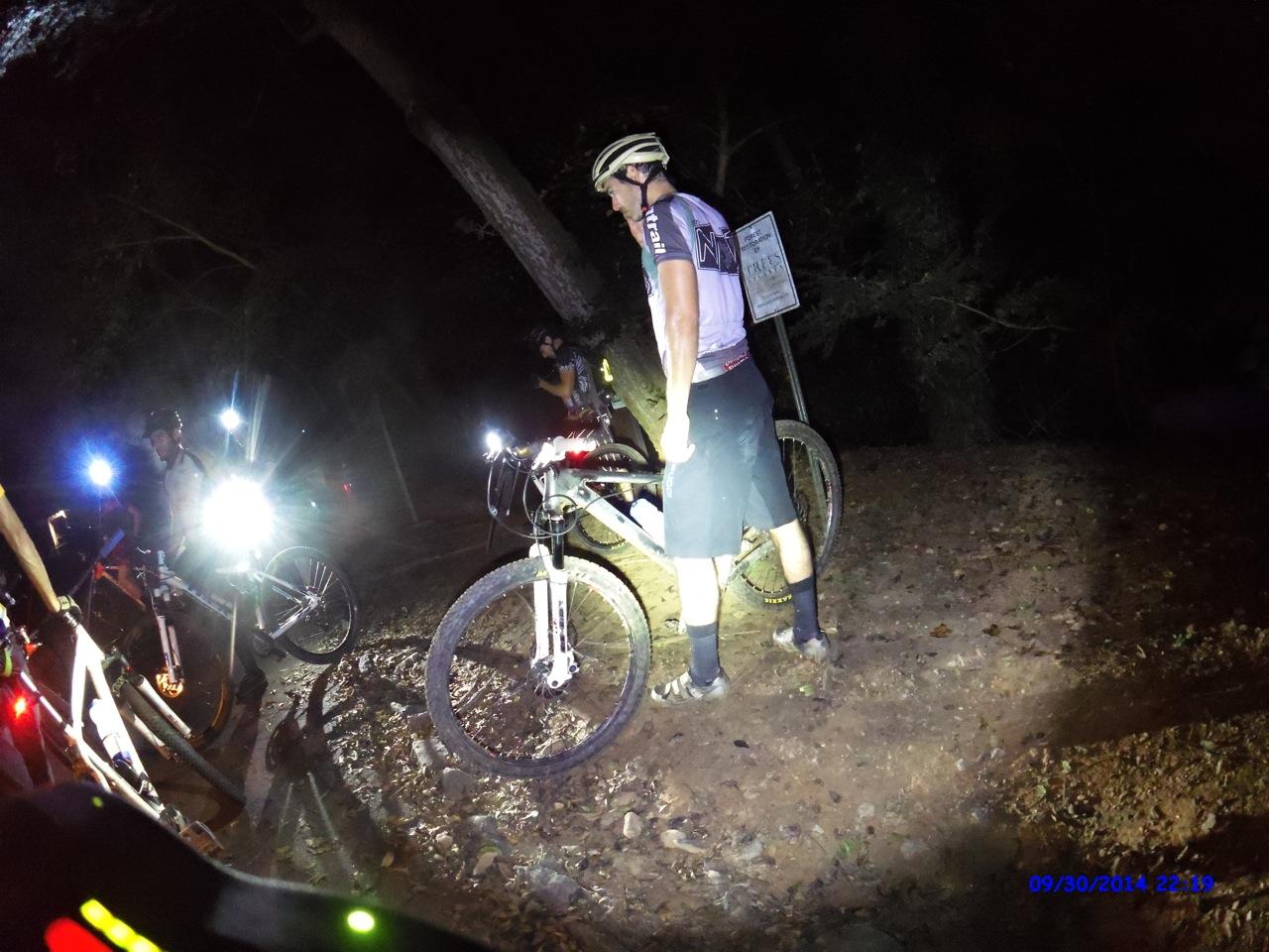 A group of mountain bikers gathered in a dimly lit forest at night. Several cyclists are visible, equipped with helmets and bicycles, while bright lights illuminate the scene, creating a dramatic contrast against the dark surroundings. One rider stands beside their bike, focusing on an area marked by a sign. The ground is covered with leaves and dirt, indicating the natural terrain of the location. Morningside Nature Preserve mountain bike trail.