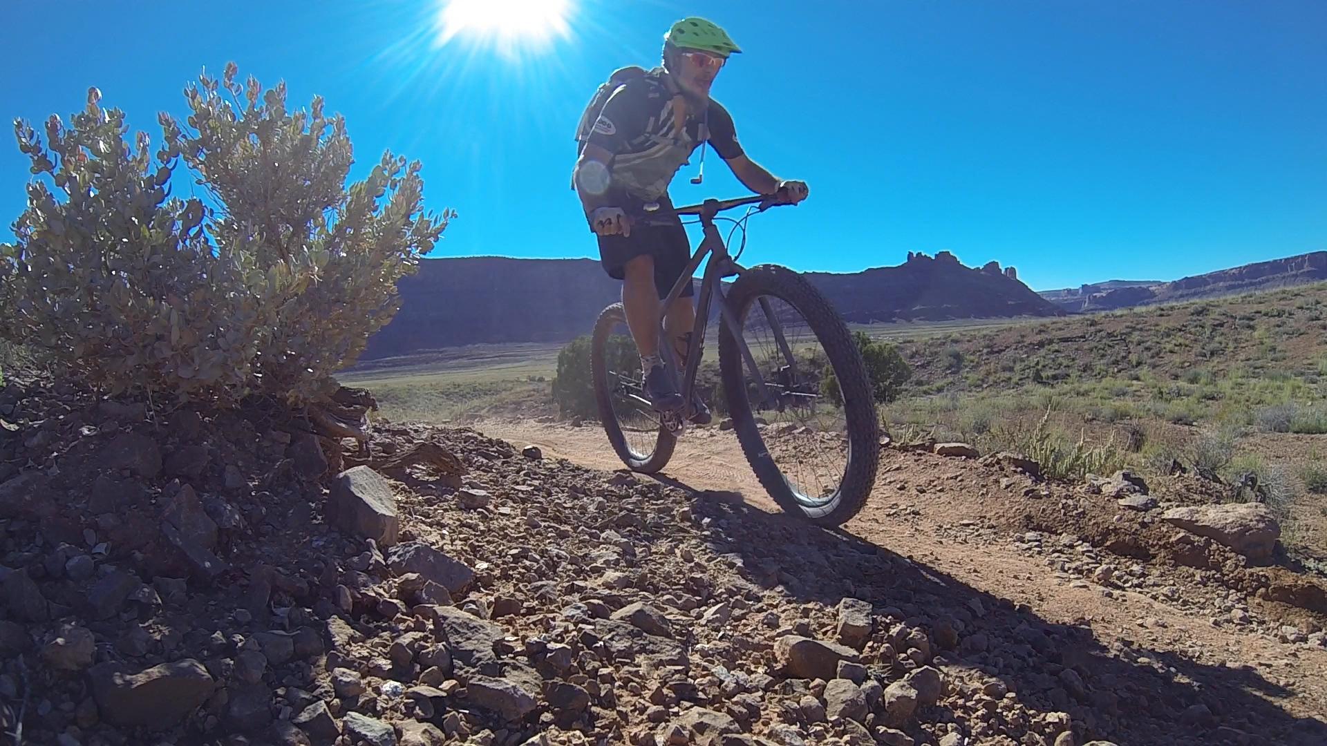A person riding a mountain bike on a rocky trail under a bright sun in a clear blue sky, with desert vegetation and cliffs in the background. Moab Brand Trails mountain bike trail.