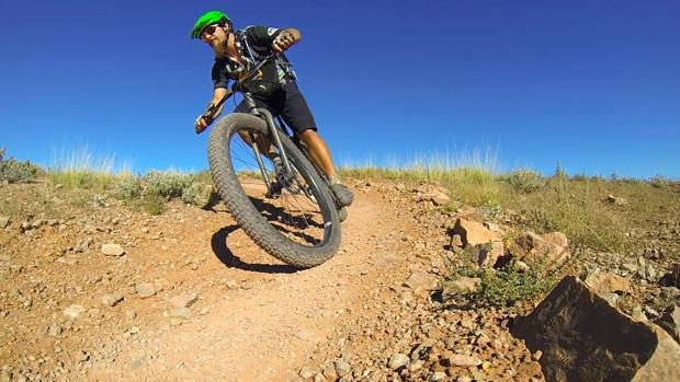 Niner ROS 9 Plus: A cyclist in a green helmet navigates a dirt trail, leaning into a turn on a mountain bike. The background features a clear blue sky and a rugged, rocky landscape with sparse vegetation.