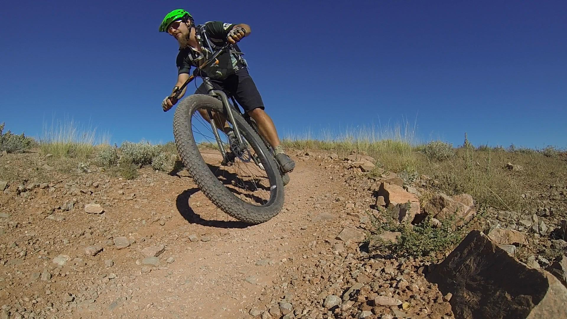 A mountain biker leans into a turn on a rocky dirt trail under a clear blue sky. He wears a green helmet and black cycling gear, showcasing an active outdoor scene. Moab Brand Trails mountain bike trail.