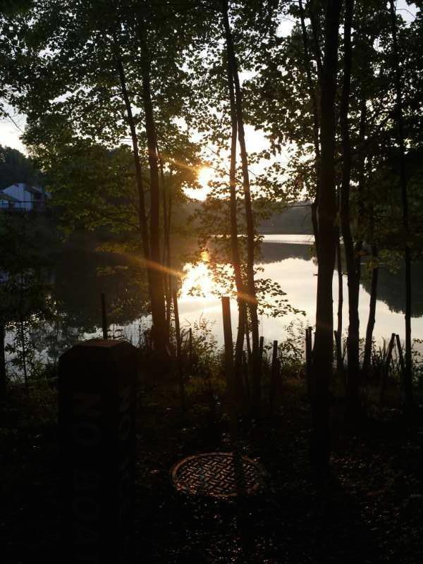 A serene landscape featuring a lake at sunset, framed by trees. The sun is partially visible through the branches, casting a warm glow on the water's surface. In the foreground, a sign labeled "No Boat" is partially obscured, adding to the tranquil atmosphere of the scene. Little Bennett Regional Park mountain bike trail.