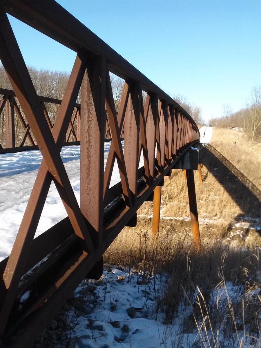 A rustic, iron bridge stretches across a snowy landscape, featuring a distinctive lattice design. The bridge casts a long shadow on the ground, which is partially covered in grass and snow. In the background, trees are visible under a clear blue sky. Raceway Woods mountain bike trail.