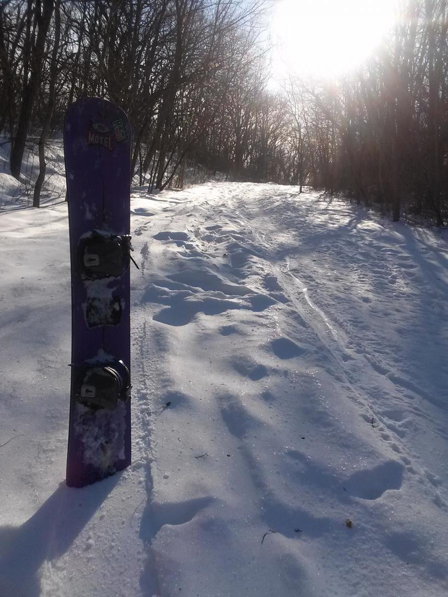 A purple snowboard stands upright in a snowy landscape, with a path leading through a forest of bare trees. The sun shines brightly in the background, creating a sparkling effect on the snow. Footprints can be seen in the snow along the path, suggesting recent activity in the area. Raceway Woods mountain bike trail.