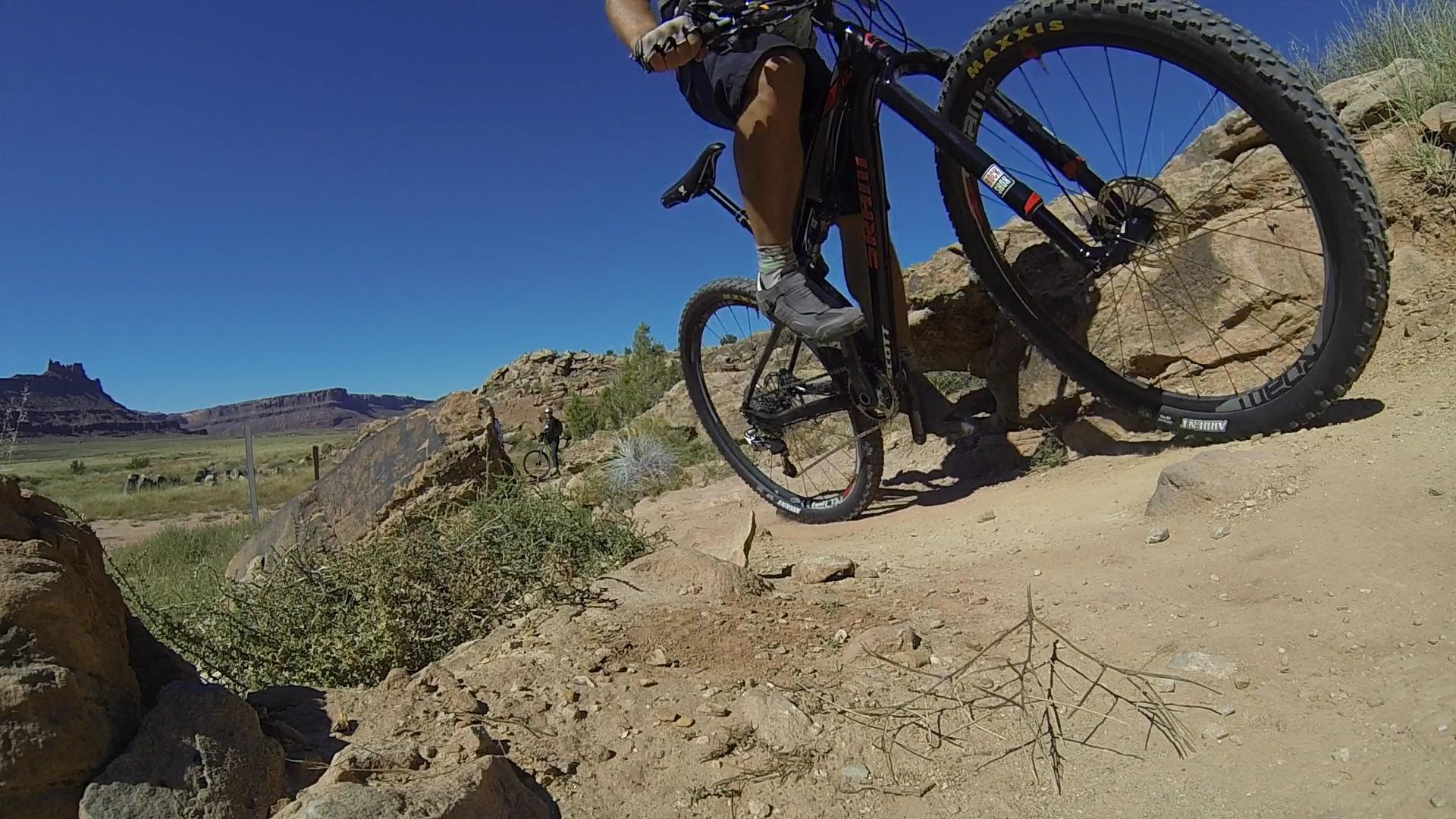 A mountain biker navigating a rocky trail with a clear blue sky overhead. The cyclist is in motion, with one foot on the pedal and the bike's front wheel lifted slightly off the ground. In the background, a scenic landscape with mountains and grass is visible, along with another cyclist in the distance. Moab Brand Trails mountain bike trail.