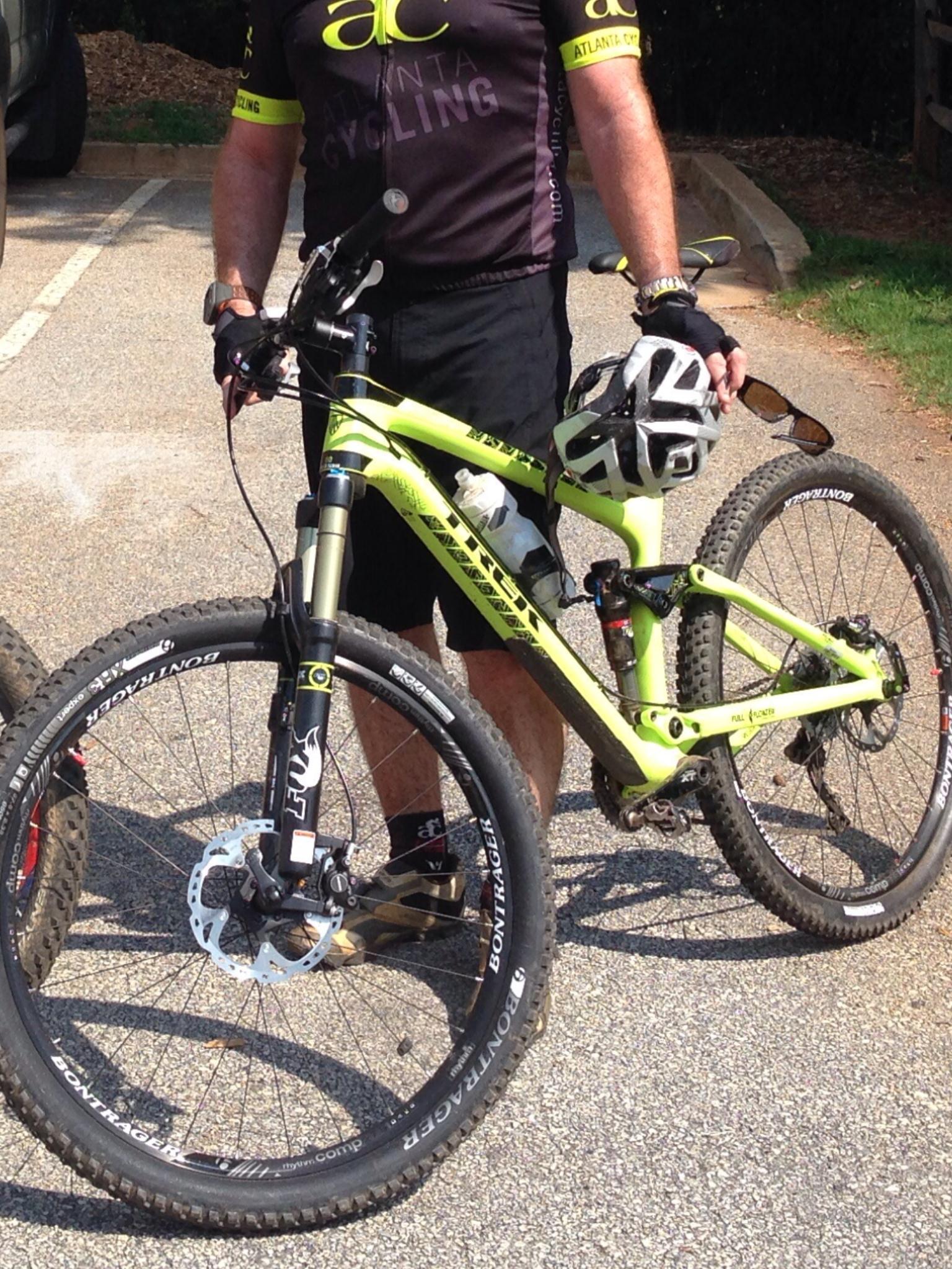 Trek Trek Fuel EX 9.8 27.5: A cyclist in a black and neon yellow cycling jersey stands next to a bright green mountain bike. The cyclist is holding a black and white helmet in one hand and has a water bottle mounted on the bike. The scene is set on a paved surface with trees in the background. The bike features large, rugged tires and a disc brake system.