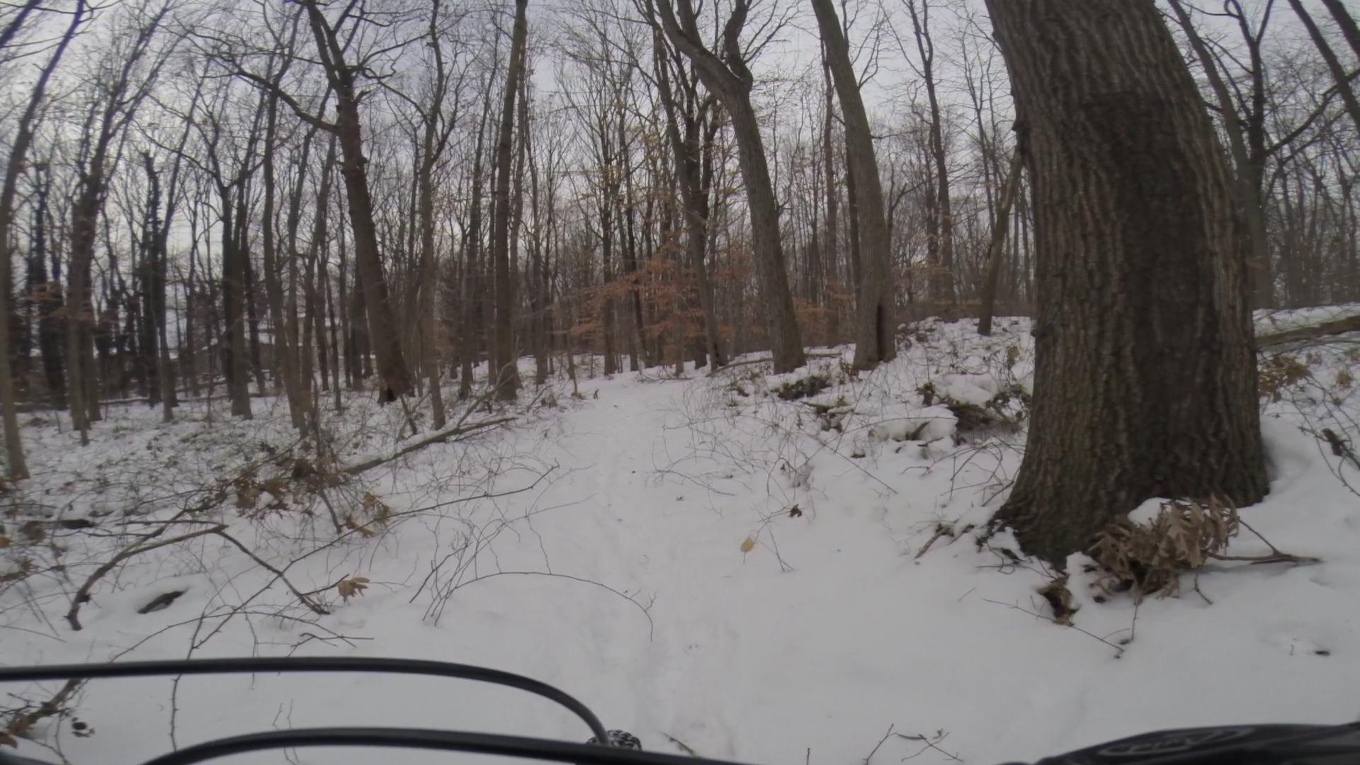 A snowy trail winding through a forest, surrounded by bare trees and patches of underbrush. The ground is covered in fresh snow, creating a peaceful winter landscape. The perspective shows part of a vehicle's handlebars in the foreground, indicating a path likely used for biking or hiking. Trails seperated by streets mountain bike trail.