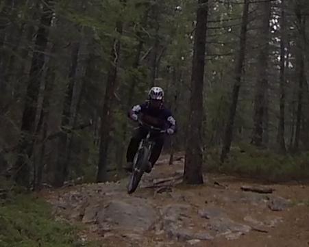 A mountain biker jumps over a rocky terrain in a forested area, surrounded by tall trees. The rider is dressed in protective gear, including a helmet, and is captured in mid-air, showcasing an action-packed moment in mountain biking. Trail 7 / 7H (Teahouse) mountain bike trail.