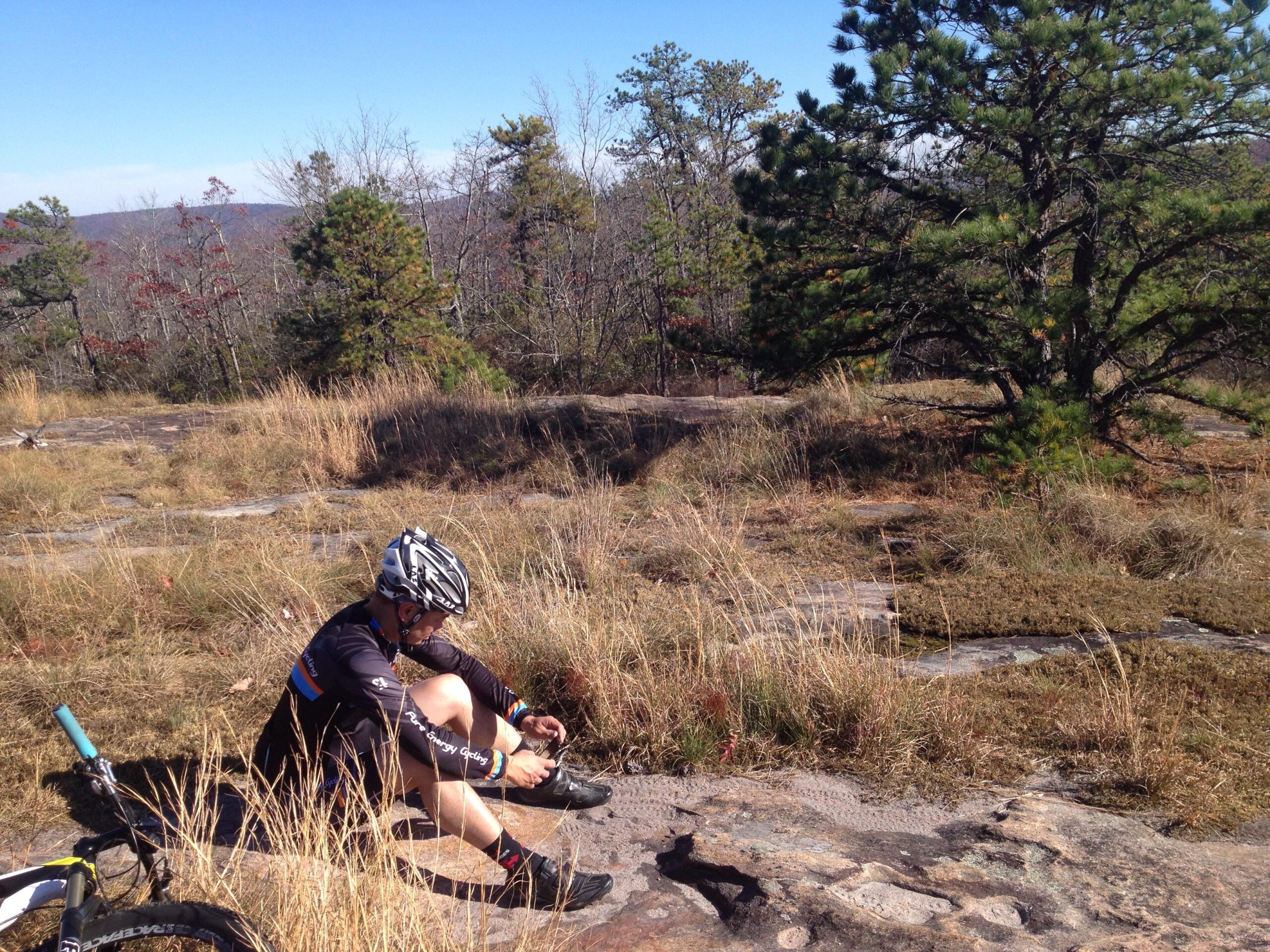 A cyclist sits on a rocky surface in a natural setting, focusing on tying his shoes. Surrounding him are patches of grass and sparse trees under a clear blue sky. A bicycle rests nearby, indicating a break during an outdoor ride. DuPont State Recreational Forest mountain bike trail.