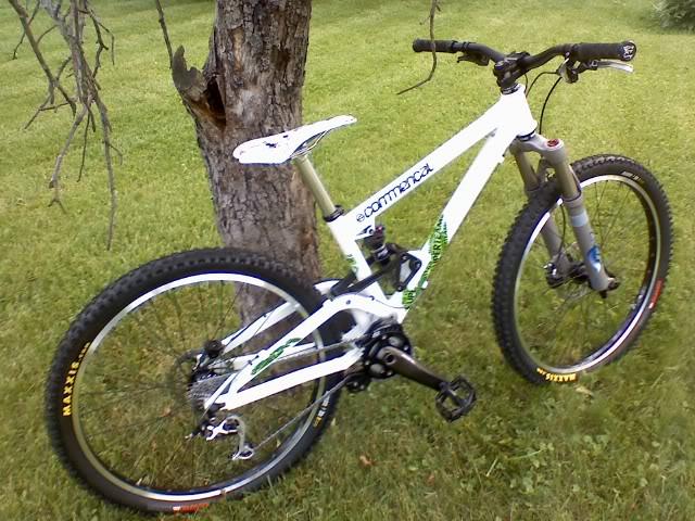 Commencal Meta HT 2: A white mountain bike leaning against a tree, positioned on a grassy area. The bike features wide tires, suspension forks, and a colorful design with green accents. The background includes a tree trunk and a clear, green lawn.