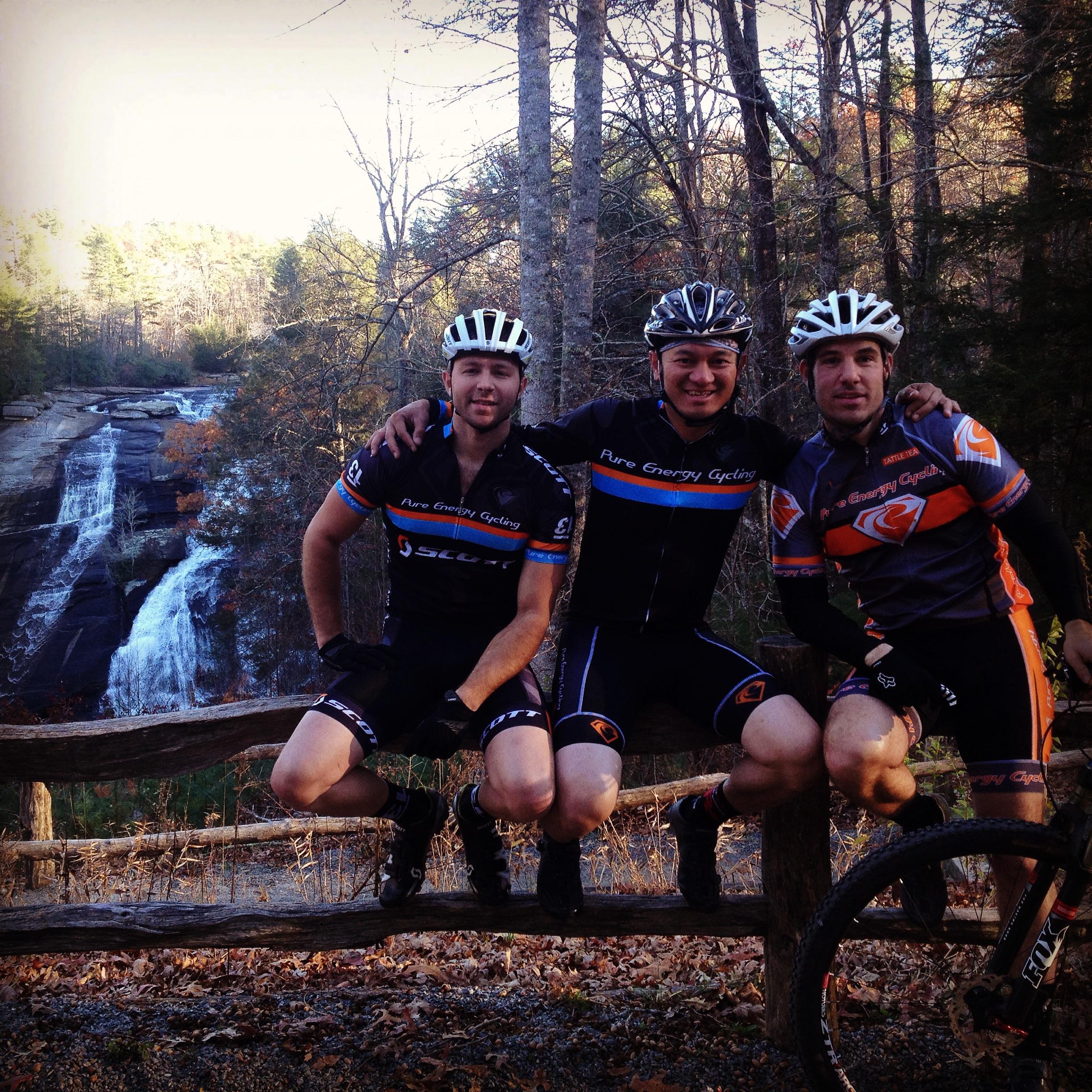 Three cyclists are posing together on a wooden fence, with a waterfall and autumn foliage in the background. They are dressed in cycling gear, featuring different branded jerseys and helmets, enjoying a break during their ride. DuPont State Recreational Forest mountain bike trail.