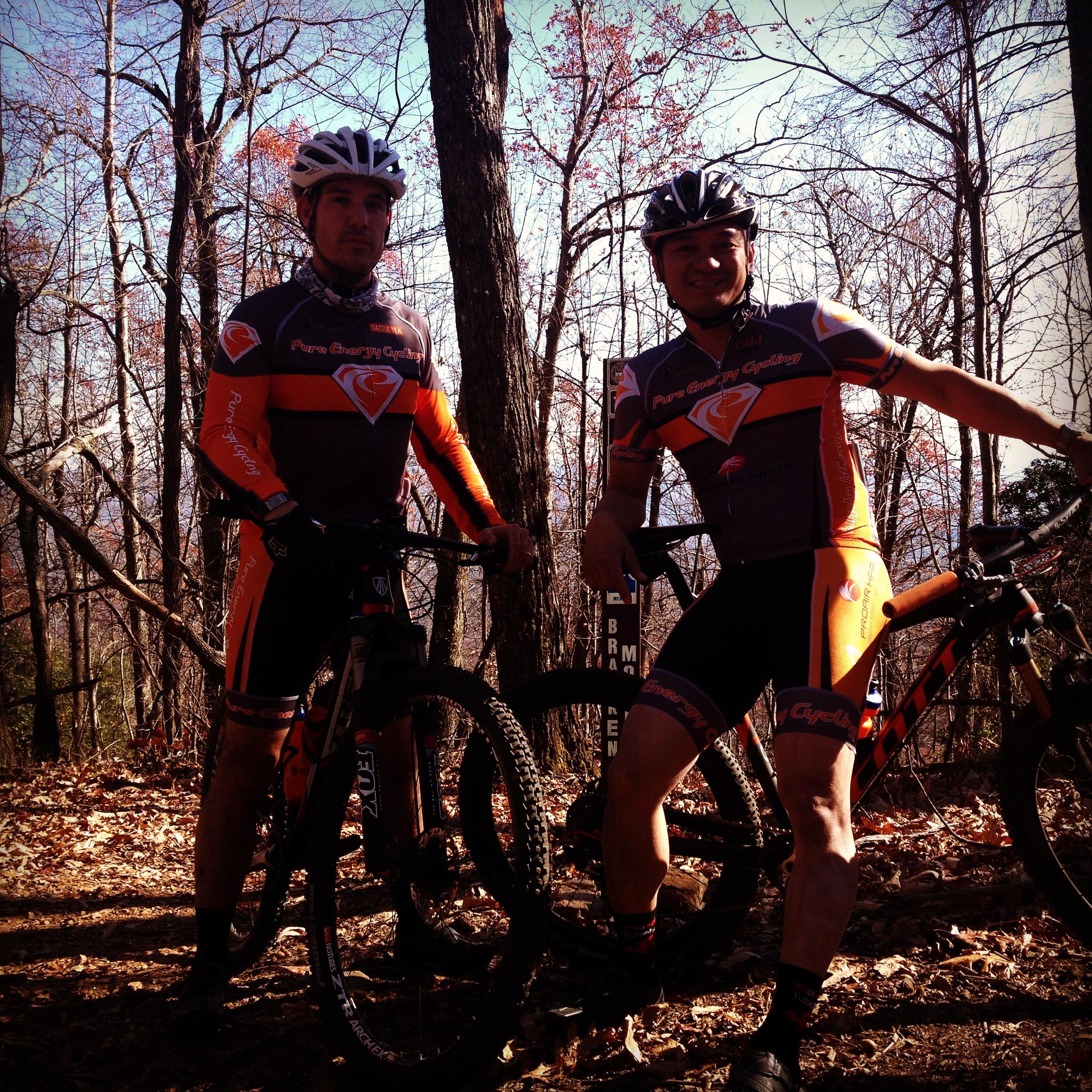 Two mountain bikers in cycling jerseys and helmets pose with their bikes in a wooded area during autumn. The background features bare trees and fallen leaves, suggesting a cool day. One rider stands confidently with his bike, while the other leans casually, both displaying smiles. Bracken Preserve mountain bike trail.