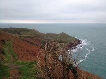 A scenic view of a coastal landscape featuring rolling hills on the left, with a rocky shore and calm ocean water to the right. The sky is overcast, creating a tranquil, moody atmosphere. Sorel Point to L'Etacq mountain bike trail.