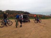 A group of four mountain bikers gathered around a parked vehicle on a dirt path. They are wearing helmets and cycling gear, with bicycles positioned nearby. The background features a scenic view of hills and a body of water, suggesting an outdoor biking adventure. Sorel Point to L'Etacq mountain bike trail.
