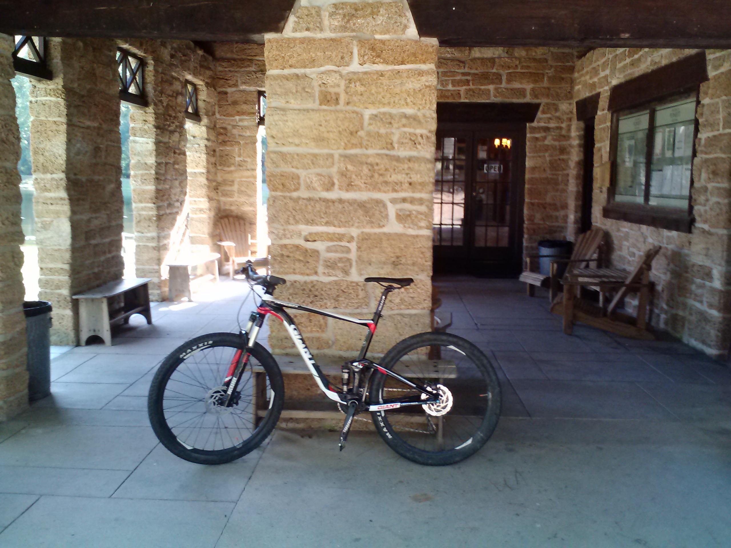 A mountain bike resting against a stone pillar in a rustic building with wooden benches and large windows. The setting is bright and includes a view of greenery outside. The Knot mountain bike trail.
