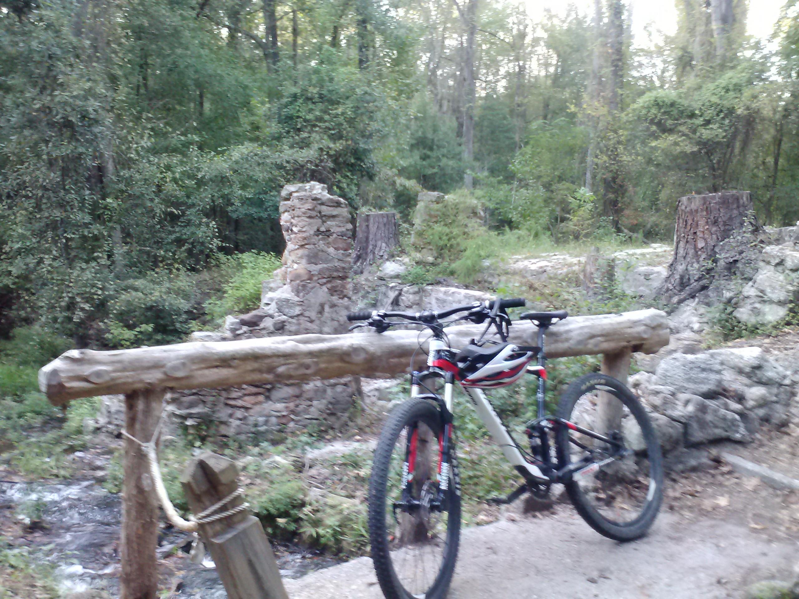 Mountain bike resting on a wooden rail near a stone ruin in a forest setting, with trees and greenery in the background. A small stream can be seen nearby, adding to the natural ambiance. The Knot mountain bike trail.