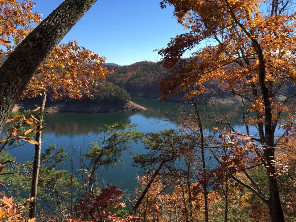 A tranquil view of a lake surrounded by colorful autumn foliage, with vibrant orange and red leaves on the trees framing the scene. The calm water reflects the clear blue sky and the rolling hills in the background, creating a peaceful nature landscape. Tsali Thompson Loop mountain bike trail.