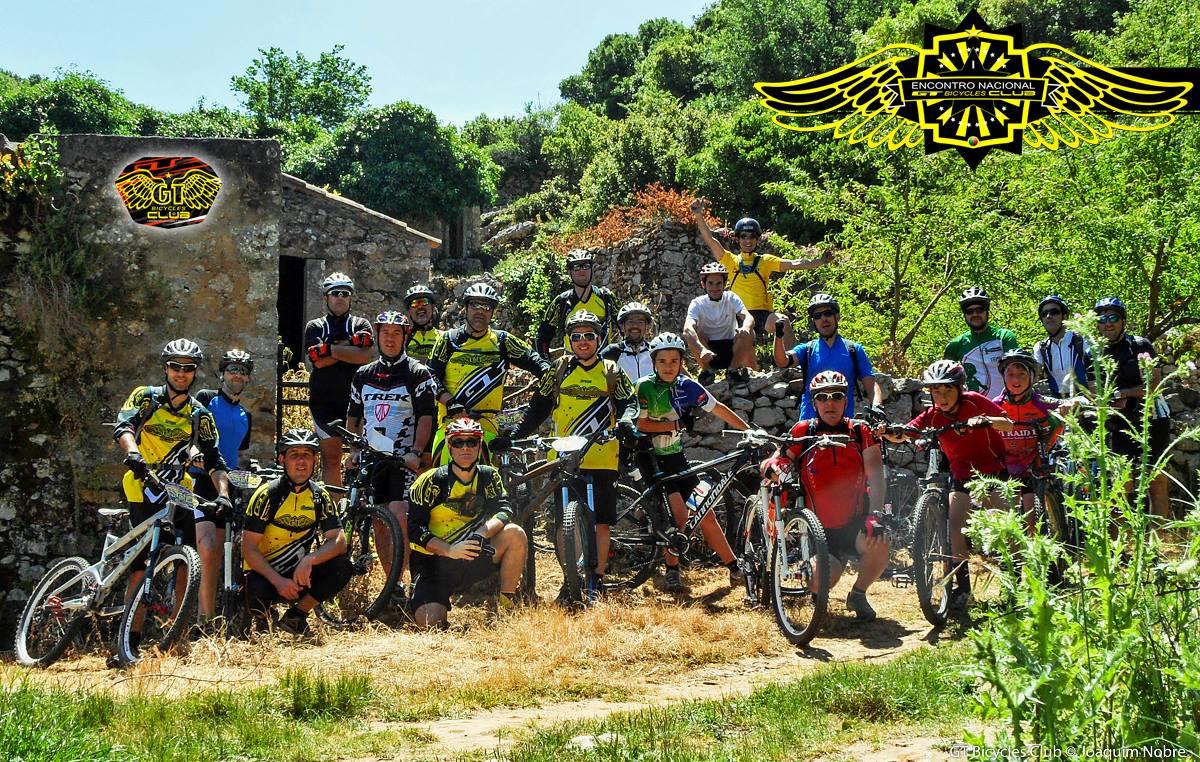 A group of mountain bikers poses for a photo in front of a rustic stone structure surrounded by greenery. The riders, wearing colorful cycling jerseys and helmets, stand with their bikes, showcasing a sense of camaraderie and enthusiasm for cycling. The scene is vibrant, capturing a sunny day in nature.