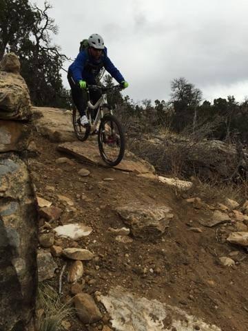A person riding a mountain bike down a rocky trail, wearing a blue jacket, black pants, and a helmet, with trees and overcast skies in the background. Oil Well Flats mountain bike trail.