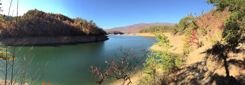 A serene panoramic view of a tranquil lake surrounded by forested hills displaying autumn foliage. The calm water reflects the clear blue sky, while the shoreline features sandy areas and various trees in shades of green and red, creating a picturesque natural landscape. Tsali Thompson Loop mountain bike trail.