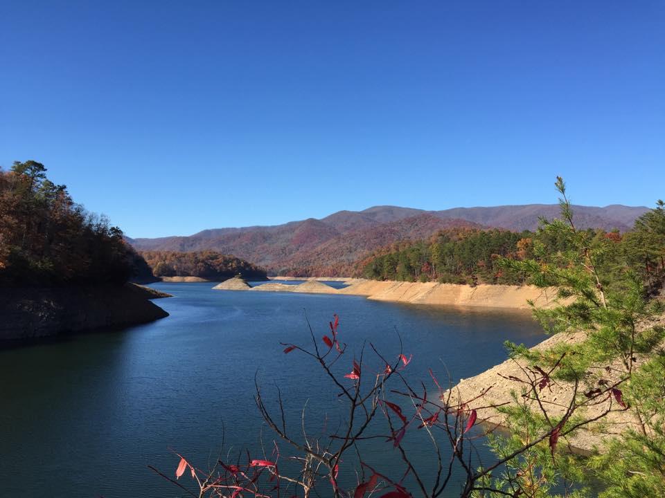A serene landscape featuring a calm lake surrounded by autumn foliage, with mountains in the background under a clear blue sky. The water reflects the vibrant colors of fall, while a few small landforms emerge from the lake, adding to the natural beauty of the scene. Green pine trees and hints of red leaves frame the foreground. Tsali Thompson Loop mountain bike trail.