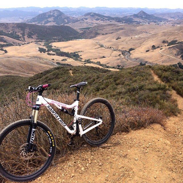 Santa Cruz 5010: A mountain bike resting on a dirt trail, overlooking a scenic landscape with rolling hills and distant mountains under a cloudy sky. The terrain is dry and brown, with patches of green vegetation.